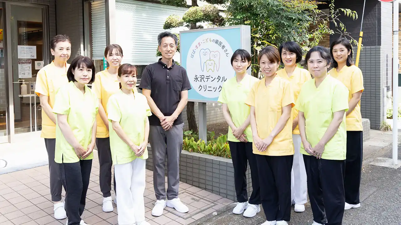 永沢デンタルクリニックの院長とスタッフが医院外の看板を挟んで並んでいる